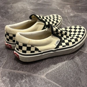 Svartvita rutiga slip-on sneakers från Vans - Klassiska slip-on sneakers från Vans med svartvitt rutmönster. Skorna har en bekväm passform och är perfekta för en avslappnad stil. Den ikoniska 'Off the Wall'-loggan syns på hälen.