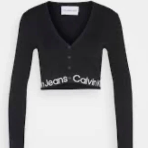 Svart croppad kofta från Calvin Klein - Snygg svart croppad kofta från Calvin Klein med v-ringning och knappar framtill. Den har långa ärmar och ett stilrent tryck med märkesnamnet längs kanten.