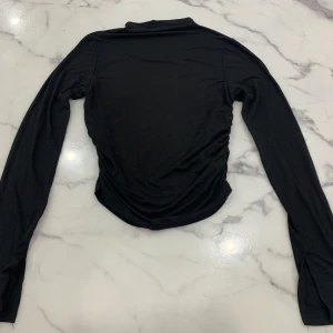 Svart långärmad topp från Gina Tricot - Säljer en svart långärmad topp från Gina Tricot i storlek M. Toppen har en stilren design med rynkade detaljer längs sidorna som ger en unik touch. Perfekt för att matcha med olika outfits.