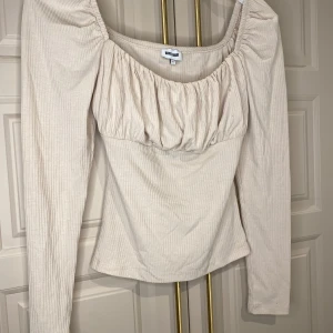 Beige långärmad topp från Bubbleroom - Säljer en stilren beige topp från Bubbleroom med långa ärmar och en vacker rynkad detalj vid bysten. Toppen har en ribbad struktur och en smickrande passform. Perfekt för både vardag och speciella tillfällen.
