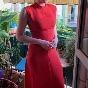 Röd ärmlös klänning - Elegant röd klänning med hög hals och ärmlös design. Perfekt för en stilren look med en figurnära passform som framhäver silhuetten. Klänningen har en klassisk och tidlös stil.