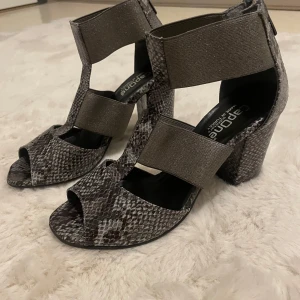 Klackar  - Snygga klackskor med ormskinnsmönster från CapOne. Skorna har en öppen tå och breda elastiska band för bekväm passform. Perfekta för en trendig look. Silver detaljer