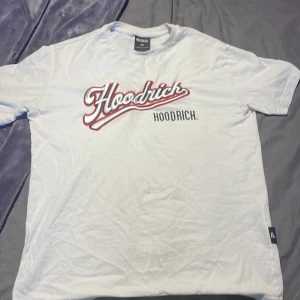 Vit t-shirt från Hoodrich - Säljer en vit t-shirt från Hoodrich med tryck både fram och bak. Framsidan har ett stort Hoodrich-tryck i rött och svart, medan baksidan har ett större tryck med texten 'Hoodrich Hood Series'. Perfekt för en avslappnad stil.
