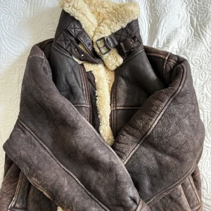 Brun Läderjacka med foder - Säljer en snygg brun läderjacka med mysigt foder. Jackan har en cool vintage look med bälte vid kragen och dragkedja framtill. Gjord av 100% äkta läder. Perfekt för kalla dagar när du vill hålla stilen. Passar perfekt för höst och vinter! med lite omvårdnad och god hänsyn blir den utmärkt!