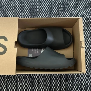 Yeezy Slides Onyx Black Strolek 44,5 - Jag säljer mina slides för att de inte  passar längre och de är knappt använda.  Slidesen är i storlek 48,5, men de har krympt när de har varit i solen. Så de passar bra till storlek 44,5-45. Hör dig om du undrar på något!