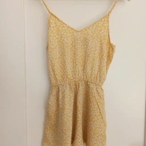 Gul blommig jumpsuit från H&M Divided - Säljer en superfin gul jumpsuit från H&M Divided i storlek XXS. Klänningen har ett fint blommönster i vitt och är perfekt för sommaren. Den är ärmlös med smala axelband och har en elastisk midja som ger en fin passform. Materialet känns lätt och luftigt, vilket gör den idealisk för varma dagar. Perfekt för både vardag och fest!🌟finns en minimal blå fläck på framsidan vid midjan, men syns endast om man står nära.