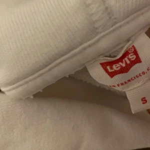 Vit hoodie från Levi's - Säljer en snygg vit hoodie från Levi's i storlek S. Den har en klassisk röd Levi's-logga på bröstet och är perfekt för en casual look. Hoodien är i mjukt material och har en bekväm passform med dragsko i huvan.