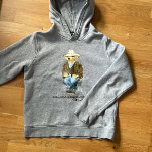Grå hoodie från Ralph Lauren med nallemotiv - Säljer en superfin grå hoodie från Ralph Lauren med ett gulligt nallemotiv på framsidan. Hoodien är i storlek xs och är perfekt för höst och vinter. Den är gjord av mjukt och bekvämt material, perfekt för att hålla dig varm och mysig. Passar både till vardags och för avslappnade tillfällen. Köp den nu eller ge ett bud! 🐻