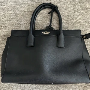 Svart handväska från Kate Spade - Säljer en stilren och elegant svart handväska från Kate Spade. Väskan är i skinn och har en klassisk design med två handtag. Insidan är randig och har gott om plats för alla dina nödvändigheter. Perfekt för både vardag och fest!