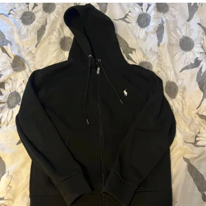Svart hoodie från Polo Ralph Lauren - Säljer en klassisk svart hoodie från Polo Ralph Lauren med dragkedja och huva. Den har en liten broderad logga på bröstet och praktiska fickor framtill. Perfekt för en avslappnad stil.
