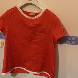 Röd t-shirt från Moncler - Snygg röd t-shirt från Moncler med vit kant vid hals och ärmslut. Den har en liten bröstficka och Moncler-logga på ärmen.  Tillhörande galge ingår!