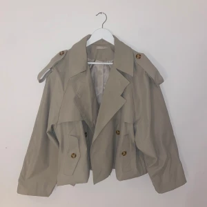Beige trenchcoat med knappar - Snygg beige trenchcoat med dubbelknäppning och axelklaffar. Jackan har en croppad design och stora knappar i brunt. Perfekt för en stilren look.