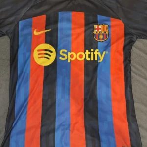 Barca tröja  - Snygg FC Barcelona tröja med klassiska blå och röda ränder. Tröjan har korta ärmar och är prydd med Nike-logga och Spotify-sponsor på framsidan. Perfekt för alla Barça-fans!