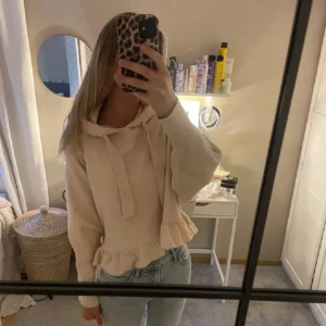 Beige hoodie med volangdetalj - Säljer en söt beige hoodie med volangdetalj i nederkant. Tröjan har långa ärmar och en mysig huva med knytband. Perfekt för en avslappnad stil.