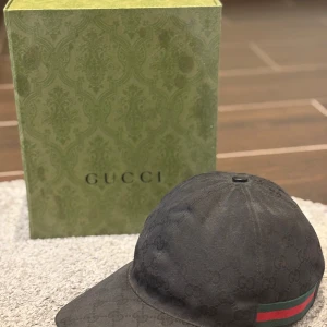 Svart keps från Gucci - Snygg svart keps från Gucci med diskret mönster och klassiska gröna och röda ränder på sidan. Perfekt för en stilren look. Allt finns med i paketet                                             Ordinare pris: 5500kr mitt pris 3000kr