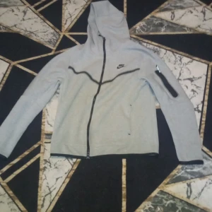 Grå huvtröja från Nike - Säljer en grå huvtröja från Nike med dragkedja och lång ärm. Tröjan har en praktisk ficka med dragkedja på ärmen och en klassisk Nike-logga på bröstet. Perfekt för en sportig look eller avslappnade dagar.