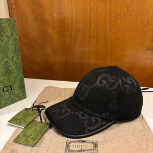 Svarta Jumbo Gucci keps - Snygg svart keps från Gucci med det ikoniska GG-mönstret. Kepsen har en justerbar rem baktill för perfekt passform. Tillverkad i ett slitstarkt material som ger en lyxig känsla. Perfekt för att ge din outfit en exklusiv touch.