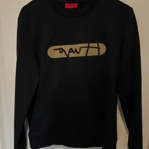 Svart sweatshirt från Hugo Boss - Snygg svart sweatshirt från Hugo Boss med ett unikt tryck i beige på framsidan. Tröjan har långa ärmar och en klassisk rund halsringning. Säljer den pågrund av att den är för liten för mig. Om ni kommer med frågor eller funderingar om priset, måtten osv så är det bara att höra av sig.