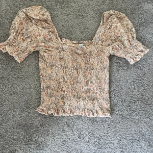 Blommig topp från & Other Stories - Säljer en söt blommig topp från & Other Stories i beige och orange toner. Toppen har korta puffärmar och en smockad design med volangdetaljer vid ärmslut och nederkant. Perfekt för en somrig look!
