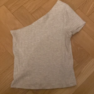 Grå ribbad oneshoulder topp från Gina Tricot - Säljer en stilren grå ribbad oneshoulder topp från Gina Tricot. Perfekt för en trendig look med sin asymmetriska design och korta ärm. Passar utmärkt till både jeans och kjol.