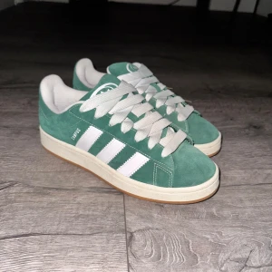 Adidas Campus 00s - Snygga gröna Adidas Campus sneakers med vita detaljer och klassiska tre ränder på sidan. Skorna har en gummisula och snörning framtill. Perfekta för en avslappnad stil. Använd endast 1-2 gånger