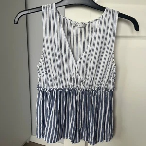 Randig ärmlös topp från ONLY - Snygg ärmlös topp från ONLY med blå och vita ränder. Toppen har en v-ringad design och en luftig passform med en elastisk midja som ger en fin siluett.