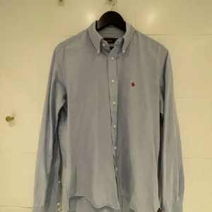 Ljusblå skjorta med broderad detalj - Snygg ljusblå skjorta med en liten röd broderad detalj på bröstet. Skjortan har långa ärmar och knappar framtill samt vid kragen. Perfekt för en stilren look.