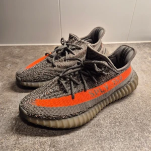 Adidas Yeezy Boost 350 V2 i grå och orange - Säljer ett par Adidas Yeezy Boost 350 V2 i grått och orange. Skorna har en stickad ovandel med snörning och en karakteristisk rand i orange med texten 'SPLY-350'. Använda 1 ggr, som Nya