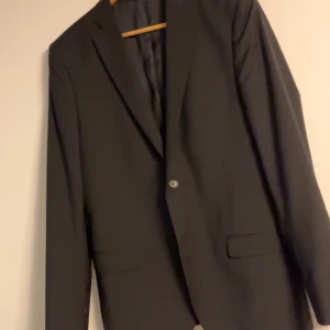 Svart kavaj från J. Lindeberg - Elegant svart kavaj från J. Lindeberg med klassisk design. Kavajen har en enkelknäppning med stilrena knappar och är fodrad för extra komfort. Perfekt för formella tillfällen. Den är sparsamt använd samt i nyskick, fickorna är inte sprättade.
