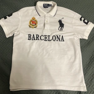 Vit pikétröja från Ralph Lauren - Säljer en vit pikétröja från Ralph Lauren med broderad logga och texten 'Barcelona' på framsidan. Tröjan har en klassisk krage med svarta ränder och nummerdetaljer på ärmarna och ryggen. Perfekt för en stilren look!