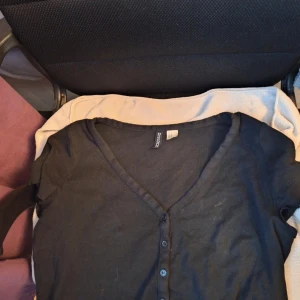 Svart T-shirt - Säljer en svart T-shirt från H&M med v-ringning och knappar framtill. Perfekt för lager-på-lager och enkel att matcha med olika outfits. Ett måste i garderoben!