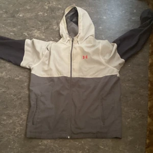 Grå och vit vindjacka från Under Armour - Säljer en grå och vit vindjacka från Under Armour med huva och dragkedja. Jackan har långa ärmar och en kontrasterande logotyp i rött på bröstet. Perfekt för blåsiga dagar!
