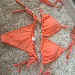 Korallfärgad bikini - Snygg korallfärgad orange rosa bikini med knytband i både över- och underdel. Triangelformad topp och klassiska bikinitrosor. Bikinin är köpt i Thailand och finns inte i Sverige!