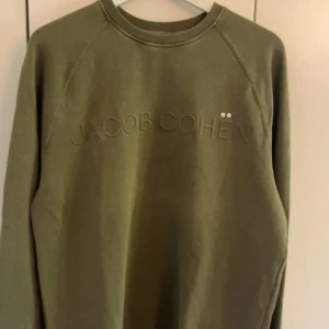 Olivgrön sweatshirt från Jacob Cohën - Säljer en stilren olivgrön sweatshirt från Jacob Cohën. Tröjan har en diskret broderad logga på bröstet och är tillverkad i mjuk bomull. Perfekt för en avslappnad look.