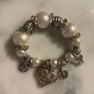 Pärlarmband med silverberlocker - Charmigt armband med vita pärlor och silverdetaljer. Armbandet har flera berlocker, inklusive ett hjärta, en fredssymbol och en stjärna. Perfekt för att ge en elegant touch till din stil.