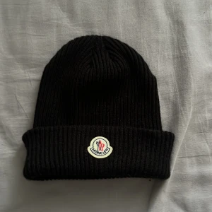 Svart ribbad mössa från Moncler - Snygg svart ribbad mössa från Moncler med uppvikt kant och det klassiska Moncler-emblemet framtill. Perfekt för kyliga dagar och ger en stilren look.