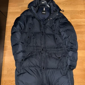 Svart dunjacka från Boss - Säljer en stilren marinblå dunjacka från Boss som är för stor för mig. Jackan har en hög krage och en insida på huvan med monogram. Den är utrustad med både dragkedja och knappar framtill samt flera praktiska fickor. Perfekt för kyliga dagar.