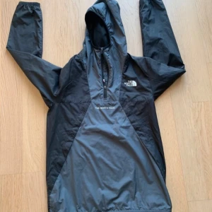 Svart vindjacka från The North Face - Säljer en svart vindjacka från The North Face i storlek XL för ungdom/junior. Jackan har en dragkedja framtill och en justerbar huva. Perfekt för blåsiga dagar och utomhusaktiviteter.