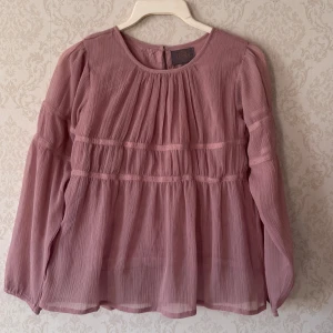 Rosa blus från Creamie - (145) - Säljer en söt rosa blus från Creamie med långa ärmar och fina rynkade detaljer. Blusen har en luftig passform och är perfekt för vår och sommar. Den är gjord i ett lätt och skönt material som ger en romantisk känsla.