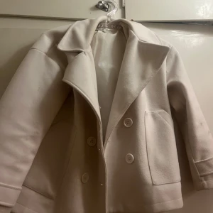 Design by si kappa - Snygg beige kappa med stora knappar och bred krage. Den har en klassisk design med två framfickor och långa ärmar. Perfekt för kyligare dagar när du vill hålla stilen.