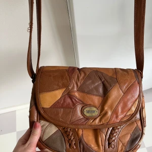 Patchwork axelväska från Carnaby - Snygg axelväska i patchwork-stil från Carnaby. Väskan är gjord av skinn i olika bruna nyanser och har en justerbar axelrem. Den har ett lock med magnetknapp och flera fack inuti för enkel organisering. Perfekt för att ge en bohemisk touch till din outfit.