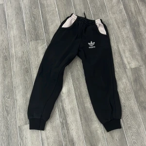 Svarta mjukisbyxor från Adidas - Säljer ett par svarta mjukisbyxor från Adidas med elastisk midja och muddar vid bensluten. Byxorna har två fickor med ljusare detaljer och Adidas-loggan på framsidan. Perfekta för en avslappnad stil.