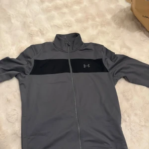 Grå träningsset från Under Armour - Säljer ett grått träningsset från Under Armour. Setet består av en långärmad tröja med dragkedja och matchande byxor. Perfekt för träning eller en avslappnad dag. Under Armour-loggan syns diskret på både tröjan och byxorna.