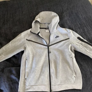 Grå hoodie från Nike - Säljer en grå hoodie från Nike med dragkedja och huva. Jackan har svarta detaljer och två fickor framtill. Perfekt för en sportig look eller avslappnade dagar.
