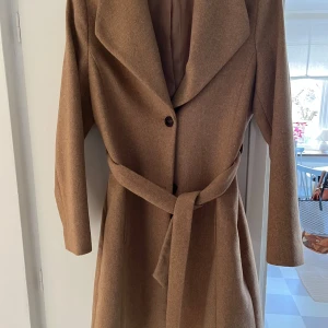 Beige kappa från Esprit - Snygg beige kappa från Esprit med knytbälte i midjan och knappar framtill. Tillverkad i en blandning av polyester och ull för en stilren look. Perfekt för höst och vår. 32% ull, resten polyester. 