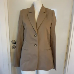 Beige ullkavaj - Snygg beige kavaj från H&M i ull. Kavajen har två knappar framtill och är helfodrad med ett slätt innerfoder. På insidan finns band där man kan spänna åt för en mer figursydd look. 
