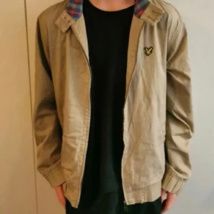 Beige vintage Lyle & Scott Harrington Jacket - Jättefin beige vintage jacka från Lyle & Scott i storlek XL.  Jackan har använts ett fåtal gånger men den ser i princip ny ut. Den enda defekten är en liten fläck bredvid dragkedjan (se bild 5), men det ser mest ut som en skugga.   Väldigt praktisk jacka som har två rymliga fickor på framsidan men även en innerficka. Dragkedja men även knappar vid kragen. Insidan är rutigt mönster i blått, rött och grönt.   Vår/Höst jacka  Köpte den för 1099