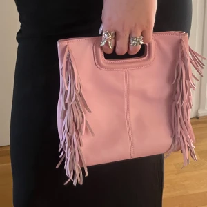 Rosa väska med fransar från Maje - Snygg rosa väska från Maje med coola fransdetaljer på sidorna. Den har ett praktiskt handtag och en avtagbar axelrem för olika stilar. Perfekt för att ge din outfit en färgklick och lite extra edge.     Köptes på hemsidan i augusti, få defekter 💞