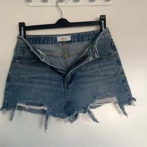 Blå jeansshorts från FGX - Snygga blå jeansshorts från FGX med slitna detaljer och fransig kant. Perfekta för en avslappnad stil. De har en klassisk femficksdesign och knappgylf. Passar perfekt för varma sommardagar.