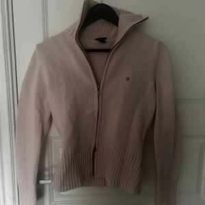 Beige stickad kofta från Gant - Säljer en stilren beige stickad kofta från Gant med dragkedja och ribbade detaljer. Perfekt för kyligare dagar med sin höga krage och långa ärmar. Den har en diskret logga på bröstet. Passar utmärkt till både jeans och kjol.